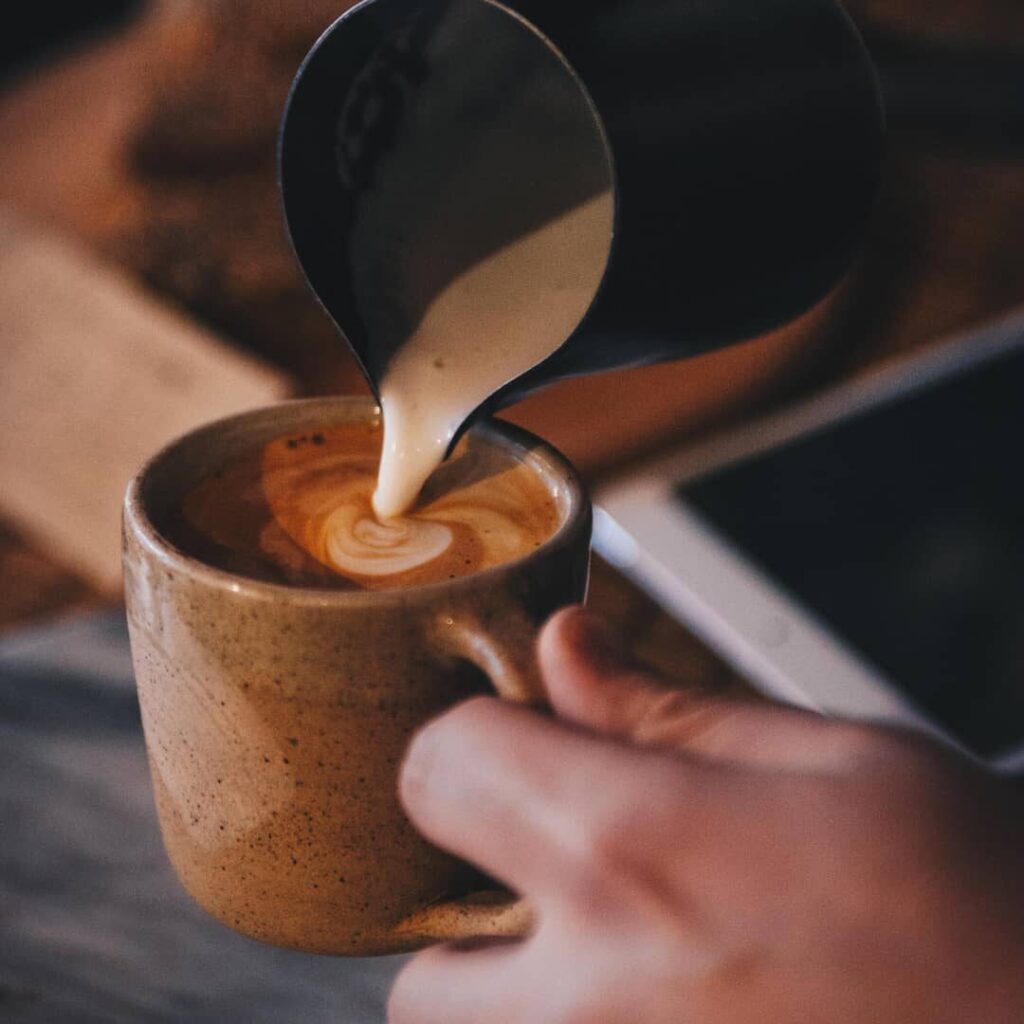 Das Bild zeigt eine Person, die gerade dabei ist, Milch in eine Tasse No Coffee zu gießen. Die Szene vermittelt eine entspannte und gemütliche Atmosphäre, ideal für den Genuss einer Tasse des koffeinfreien Kaffees. Der sanfte Strudel, der sich im Kaffee bildet, lässt darauf schließen, dass es sich um eine sorgfältig zubereitete Latte Art handelt, was die Qualität und den Genuss des No Coffee noch unterstreicht. Die rustikale Tasse und die warme Umgebung verstärken das Gefühl eines bewussten, genussvollen Moments ohne Koffein.